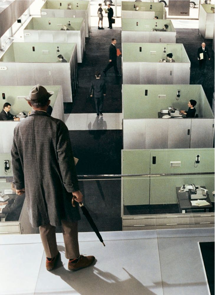 A still from the film Playtime of Jacques Tati in his Monsieur Hulot role. He is looking down on an office floor where there are a number of identical cubicles with no ceilings within which we see one person working. As with the photograph in the quoted tweet we see Tati from behind in coat and hat. No pipe this time but instead he holds an umbrella.