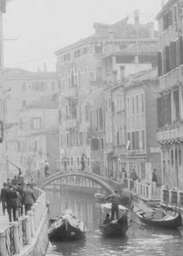 detail 📷 William Williams 
A detail from the photograph in the quoted tweet zooming in on the view down the far end of the canal where we see a number of men walking along the pavement and over a bridge, and a gondola making its way between boats. The buildings on the right side of the canal swing round to the left following the bend in the canal taking up the whole of the background apart from some sky above.