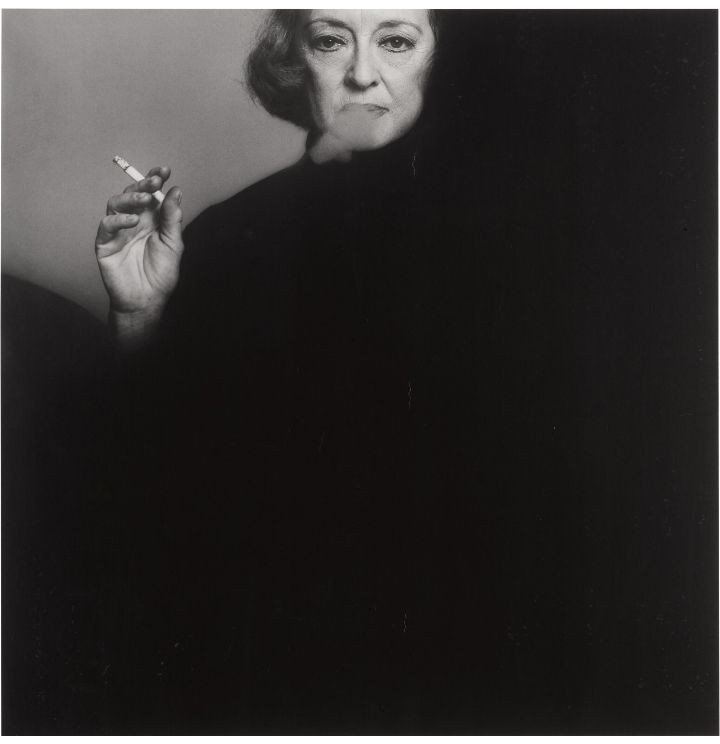 📷 Victor Skrebneski 
Black & white photograph of Bette Davis almost all black with no distinguishing features except for the top left hand corner where, against a grey background, we see Davis's hand, cigarette between fingers thumb on the butt end, her head cut off just above the eyebrows, hugged on the right hand side by the blackness, and from the mouth a puff of cigarette smoke, she looks at the camera.