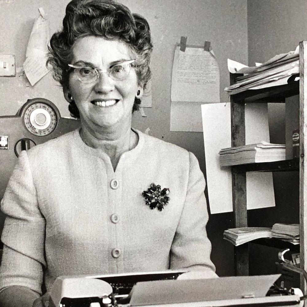 Black & white photograph of Mary Whitehouse sitting behind her typewriter, smiling at the camera.