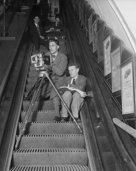 📷 unknown to me 
Black & white slightly cropped photograph of Anthony Asquith filming Underground. He is sitting to the right of an escalator with a cameraman beside him, looking out of shot down to our left. The tripod legs of the camera are on various steps across the width of the escalator. Down the right hand side framed advertisements, towards the top two further people sitting.