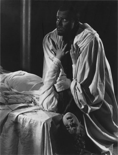 📷 Angus McBean 
Black & white photograph of Orson Welles as Othello strangling Desdemona played by Gudrun Ure. It's a staged pose for a theatrical production. Othello stands over the bed but looks away to his right, his arms quite straight, his hands around Desdemona's neck,whose head, upside down, hangs over the end of the bed looking in our direction, mouth open, her hands grasping up by his chest. Black background.