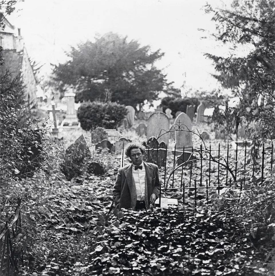 📷 John Deakin 
Black & white photograph of Dylan Thomas who appears to be standing up to his waist in the weeds of an overgrown cemetery, although this may partly be due to a trick of the angle. Behind him we see grave stones. He is wearing a jacket and shirt with a bow tie. This photograph was chosen to compare with the photograph in the quoted post of the Beatles standing in similar.