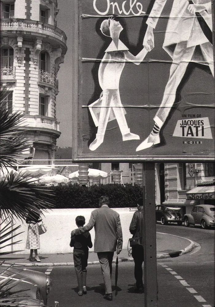 📷 Henri Cartier-Bresson 
Black & white photograph of Jacques Tati and the young Alain Becourt walking over a road ahead of us, Tati with his hand on the boy's shoulder, umbrella in his other hand. They would have just walked past, or under, a sign that we see only part of in the top right corner of the photograph. It shows a cartoon-like figure of a boy holding an adult's hand. We see the adult has an umbrella and the french word Oncle appears and the name Jacques Tati. This is a promotional poster for the film they've just made.