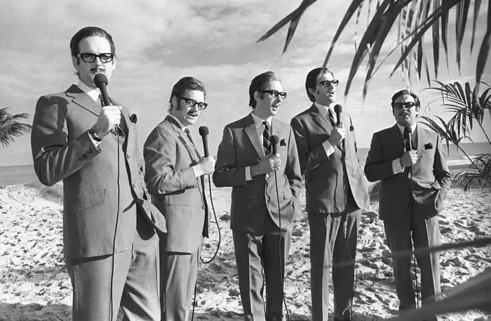 A photographic still from the Monty Python sketch Whicker Island. Five of the Monty Python crew dressed as Alan Whicker, with microphone in hand, all in a line on a sandy beach, sea in the background, palm tree appearing into shot on the right.