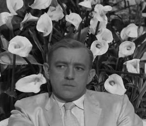 Black & white still from the film Our Man in Havana showing Alec Guinness sitting down surrounded by lilies. He looks to his right oblivious of how the arrangement looks to us. 
Photograph picked to compare with the photograph in the quoted post.