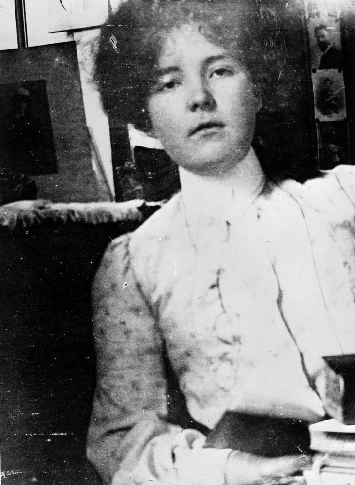 📷 Unknown photographer
Black & white photograph of Katherine Mansfield. She leans to her right into the top centre of the shot, the top of her head out of frame, she looks down at the camera, open book in her right hand which is just behind a pile of books that we get a glimpse of in the bottom right hand corner. There's a window behind her, and left and right are photographs that are difficult to make out (literary heroes perhaps, one looks like Swinburne). A chance photograph or one well worked out and casually performed.