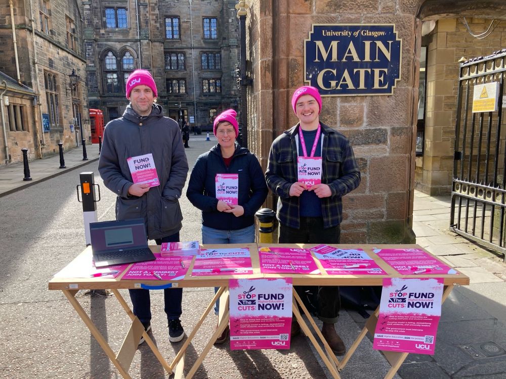 Three people in pink hats, holding flyers which say Stop the Cuts, behind a table with big pink posters and a laptop.