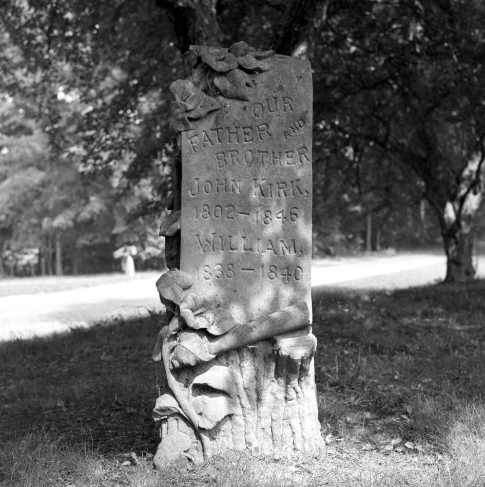 an ornate gravestone from the 1840s, sculpted to resemble a tree stump with flowers. inscribed "our father and brother john kirk 1802-1846 william 1838-1840" photographed in black and white