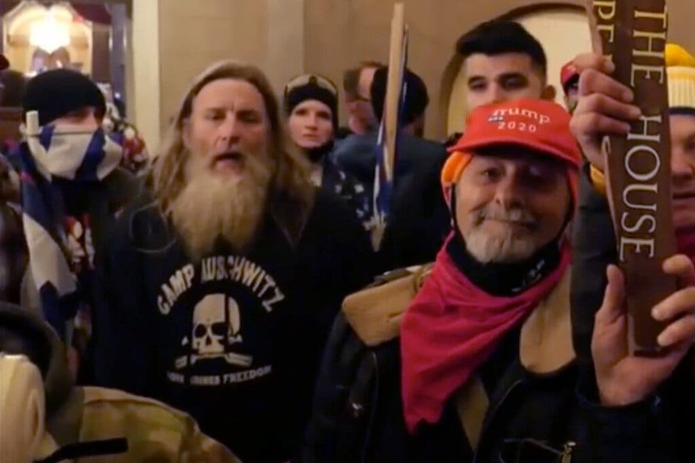 Ugly J6 domestic terrorists proudly desecrating U.S. Capitol. Man on left with disgusting scraggly beard that looks like health violation is wearing black shirt with skull & crossbones & words, "Camp Auschwitz" on top. The absolute worst dregs of humanity.
