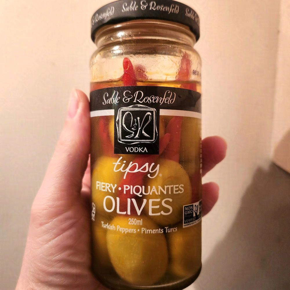 A glass bottle with a black metal top held aloft by a very white woman's hand against an off-white wall. 
The text on the bottle reads (summarized)
"Sable and Rosenfeld Tipsy Firey Olives"