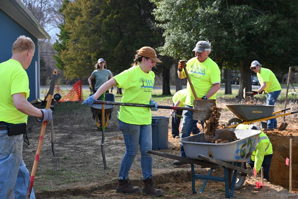 Volunteers in bright yellow shirts work together on a landscaping project, shoveling soil into a wheelbarrow near a freshly dug trench. Several people are actively using tools, while others assist or observe nearby. The scene is set outdoors beside a blue building and surrounded by trees.