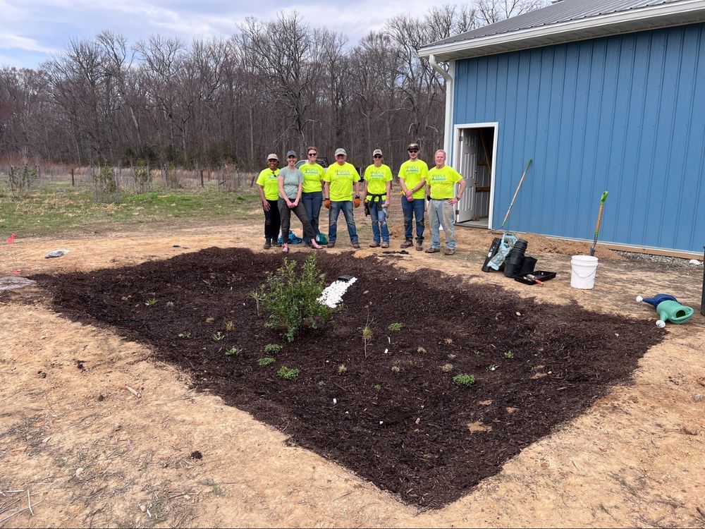 A group of eight people wearing bright yellow shirts stands smiling and posing in front of a newly planted and mulched rain garden next to a blue building. Gardening tools and supplies are scattered nearby, and a wooded area is visible in the background.
