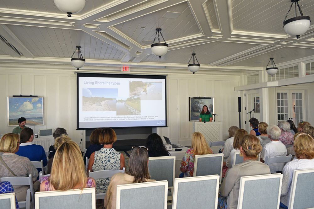 A woman speaks at a podium with a presentation on living shorelines projected beside her. People are seated in the room listening to her