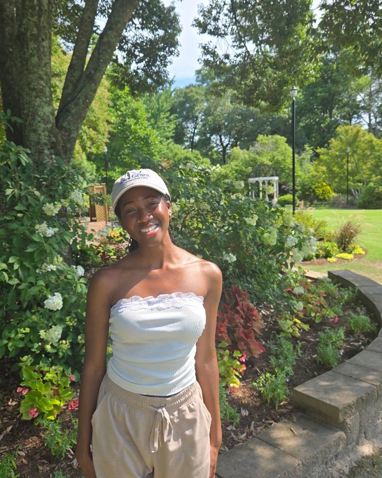 A young woman poses for a photo in an arboretum with flowers, shrubs, trees, and grass on a sunny day