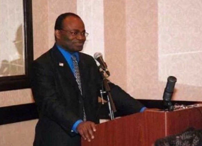 Spectacled Black man at podium with gray patterned tie, blue shirt, and black suit. He’s bracing himself on the podium with the mic up to his mouth. He’s smiling in a smug, almost mischievous way. He knows he’s about to rock his audience’s minds
