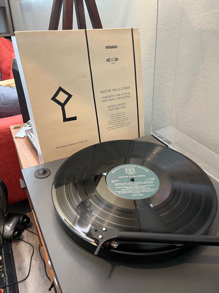 A turntable with a record playing; a Heitor Villa-Lobos LP jacket is leaning against a lamp alongside