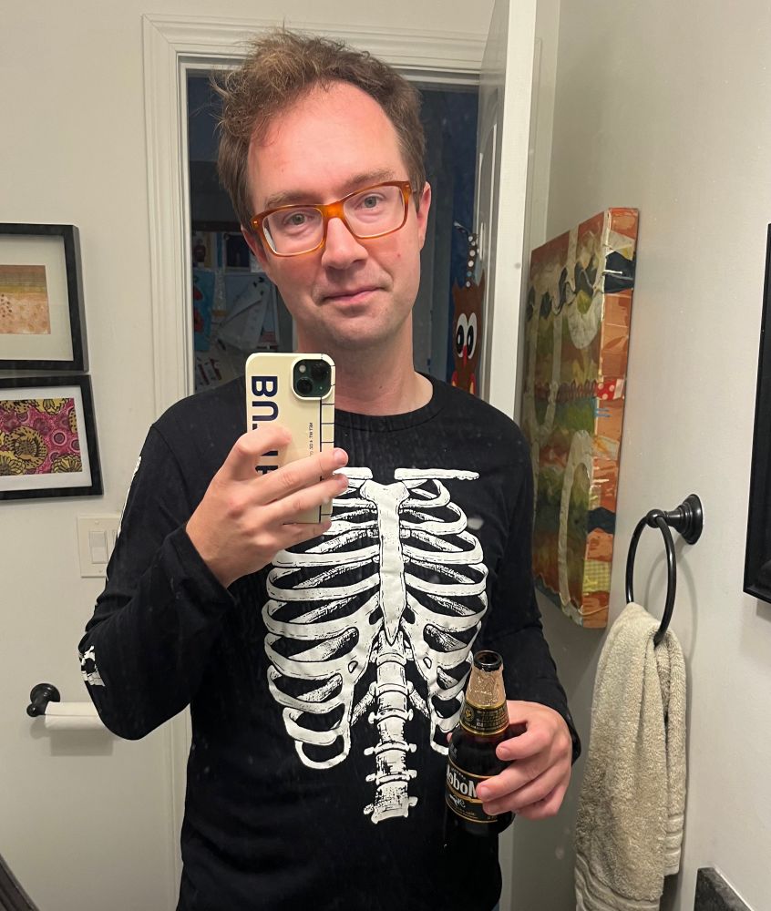 a selfie of a middle aged man in glasses with reddish brown frames, holding a beer bottle in the left hand and a phone in the other 