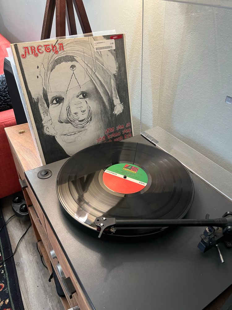 A turntable with a record playing; Aretha Franklin’s Hey Now Hey (The Other Side of the Sky) LP jacket is leaning alongside
