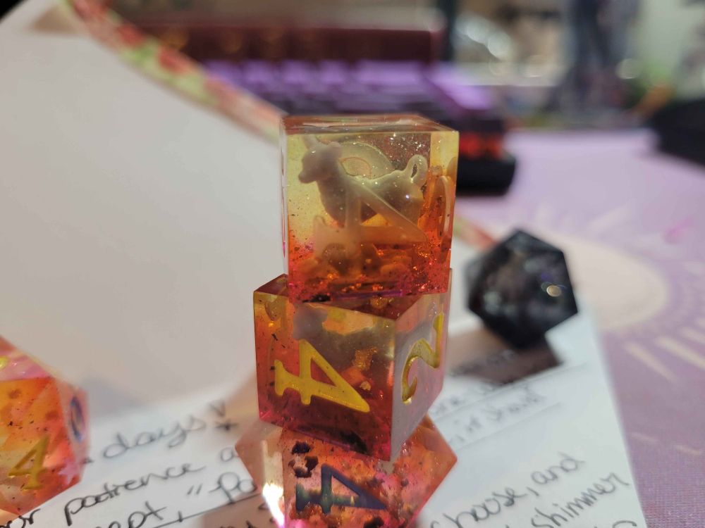 The recipients photo of the dice. the unaponted D6 is balanced on top of the painted d6, which is balanced on a d4.
Because of their excellent photography, you can see the colors diffused from the light. Bright oranges, swirls of magenta pink, and a large amount of pale yellow. The d6s have beautiful Laika on full display.