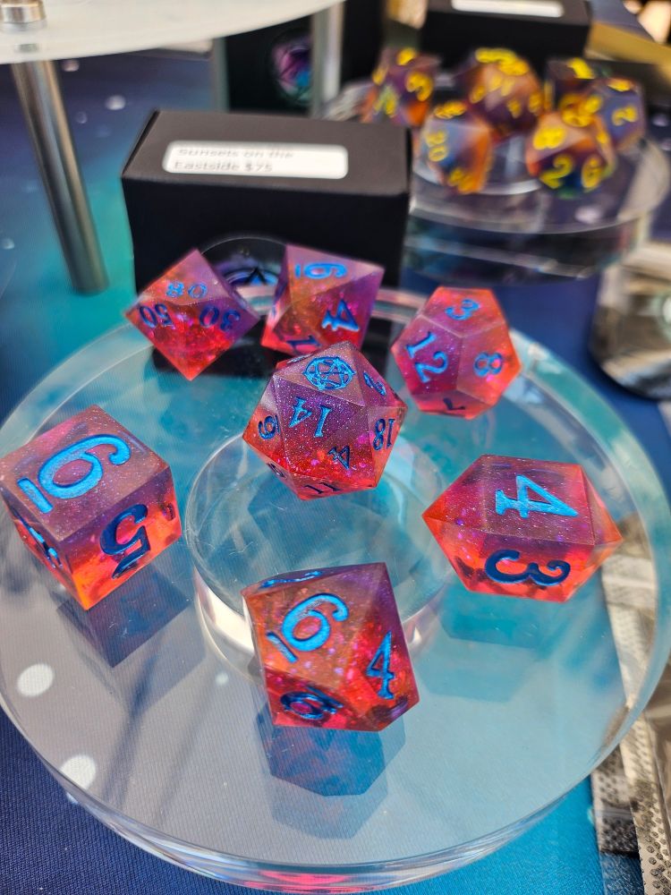 A full polyhedral set of dice, colors of deep translucent orange to purple dice with glitter and shimmery inclusions, inked with a deep reflective metallic blue, named Sunsets on the Eastside