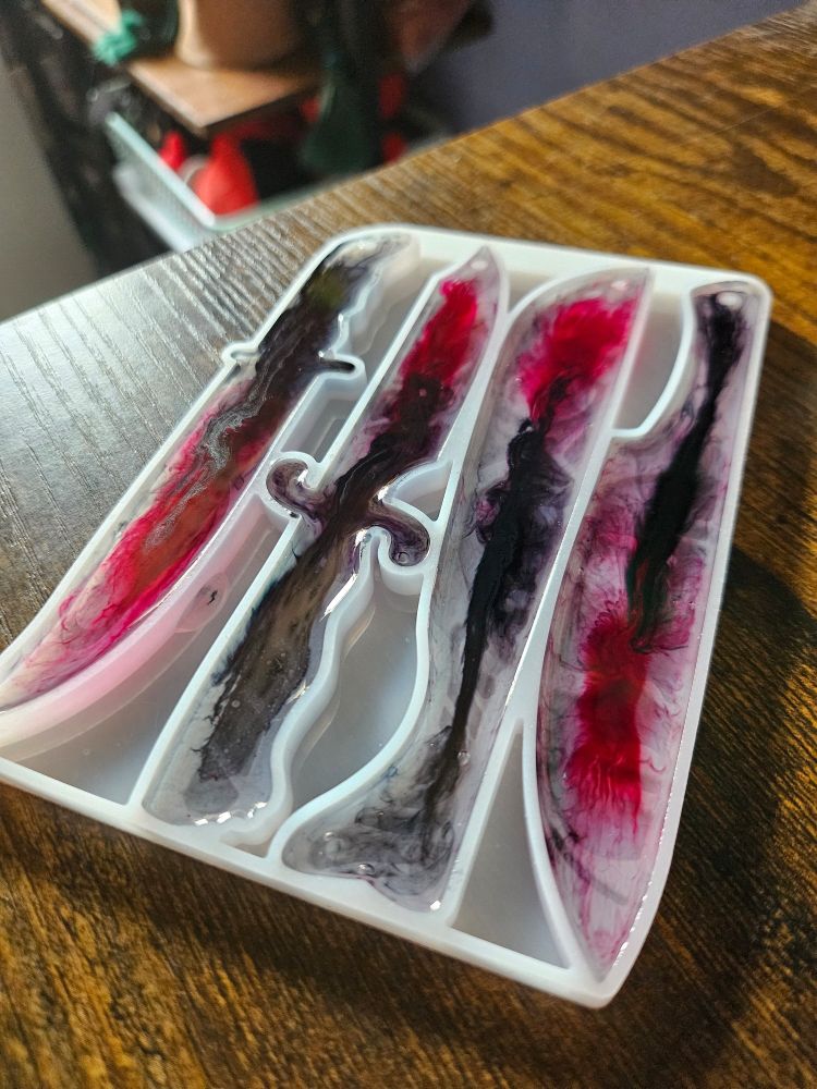 A mold of 4 bladed weapons, sitting on a wooden desk corner; resin is poured into the mold with black and red ink swirls. It's not quite done curing.
