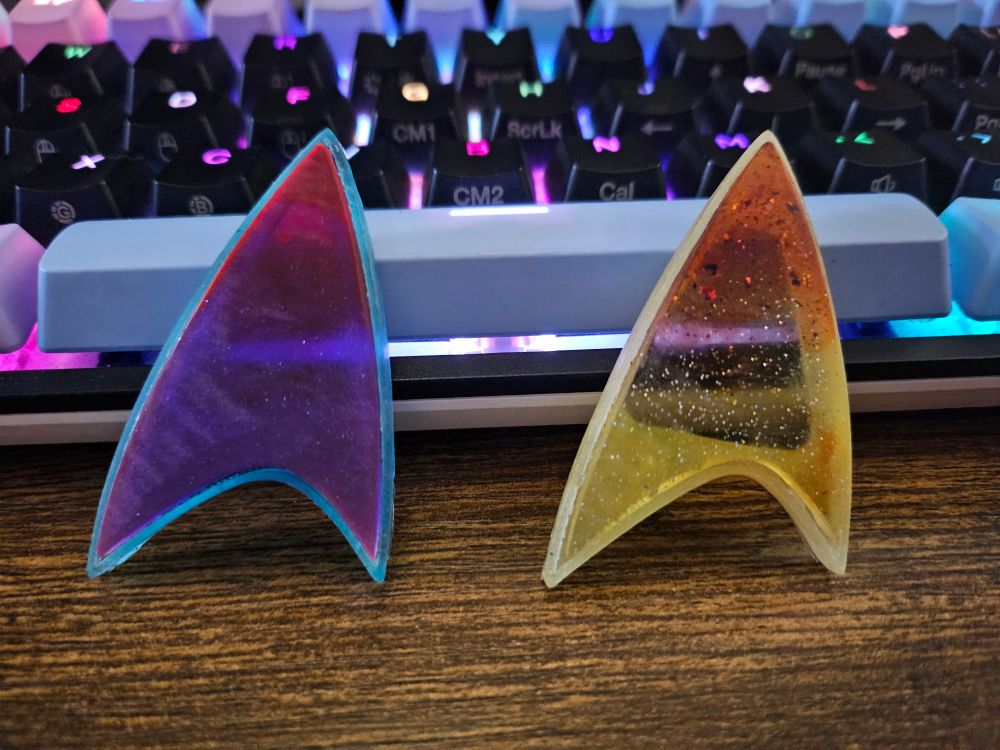 Sitting upright against a keyboard, Two resin star trek badges, one is blue outline with purple and reddish/pink for the body, and the other is white/yellow outline with a sparkly yellow and orange body. They're slightly transparent, so some of the keyboard backlighting shines through.