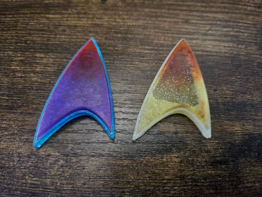 Laying flat on a desk are two resin star trek badges, one is blue outline with purple and reddish/pink for the body, and the other is white/yellow outline with a sparkly yellow and orange body. 
