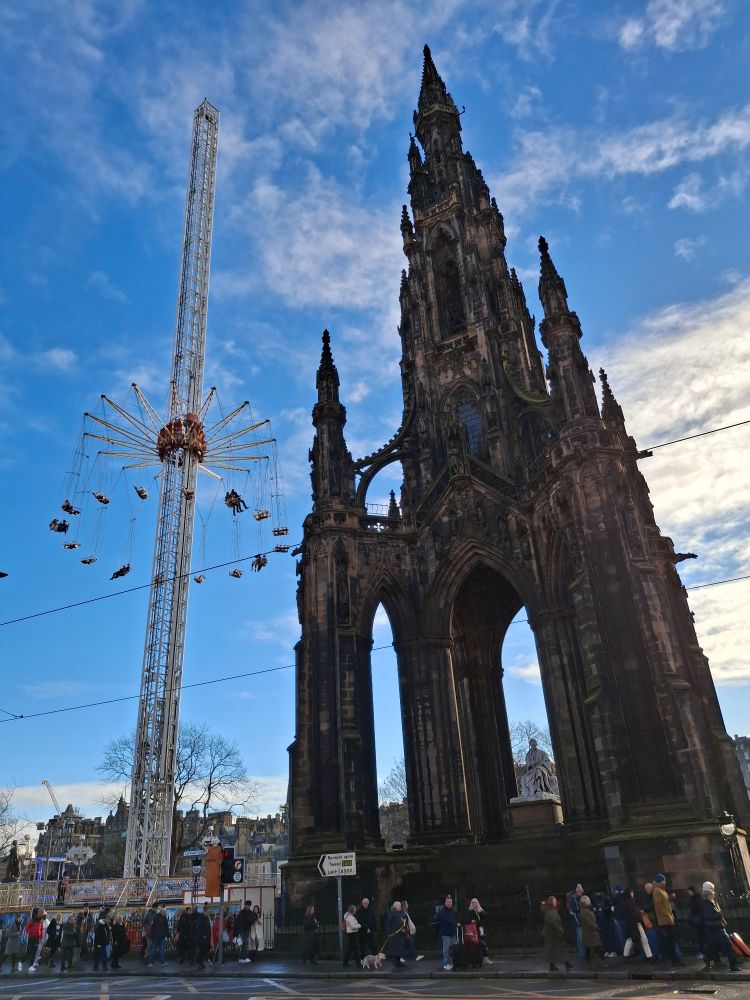 The Scott Monument in Edinburgh next to a tall modern fairground ride with a lightly clouded blue sky in the background 