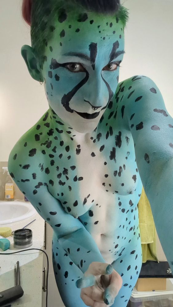 This photograph shows a woman named Pikelet with elaborate, full-body paint.

She is depicted as a stylized, colorful feline, reminiscent of a cheetah but with a fantasy color scheme. Her skin is painted a vibrant turquoise blue, which transitions into a pale, whitish-green on her face and down the center of her torso. The blue base is covered in black, irregular spots.

Her face is also fully painted, with a white "muzzle" area, black lips, and dramatic black markings around her eyes to enhance the feline look. Her hair is colored green and appears to be styled back.

Pikelet is looking at the camera with a smile and making a "peace" sign gesture with her hand. The photo appears to have been taken in a bathroom, with a sink, counter, and towel visible in the background. The overall impression is one of creative and playful body art.