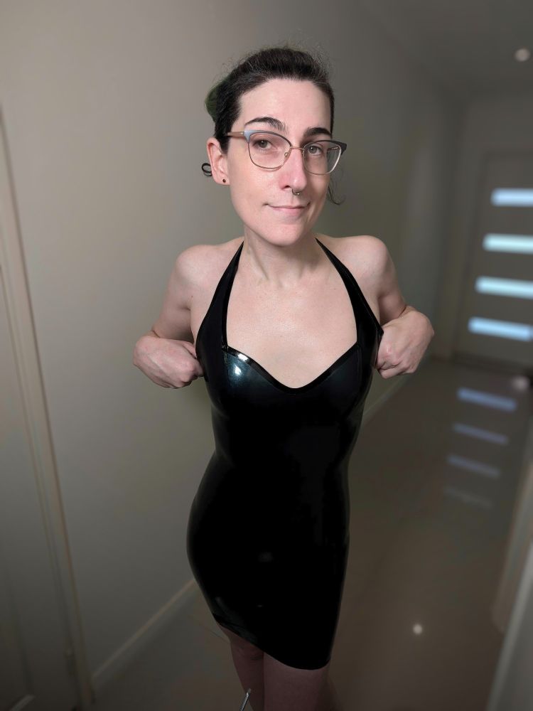 Woman with glasses and long green undercut hair which has been growing out to reveal the natural black hair wearing a latex dark blue metallic skin tight halter dress looking at the camera with a smirk and posing looking directly at the camera and leaning over slightly while pulling up on the top of the dress just below the straps.
