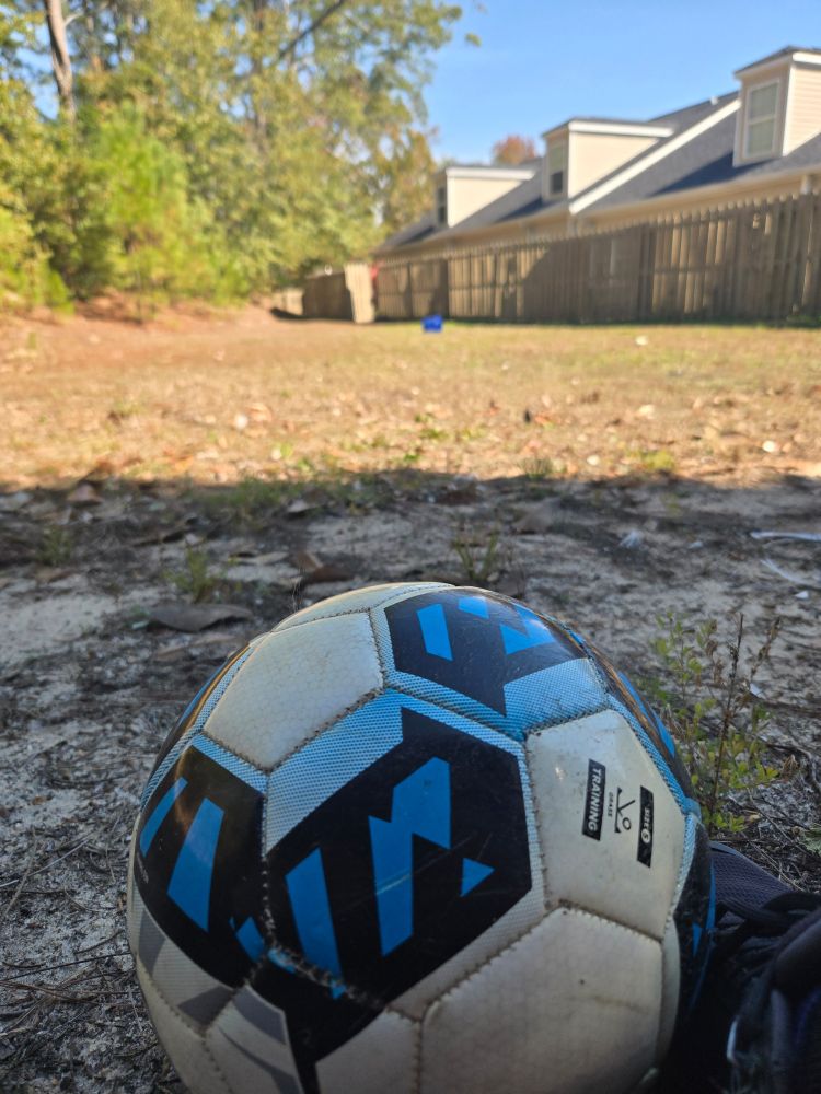 A soccar ball is in focus up cloae, with a blue Lowes bucket out of focus in the distant grass