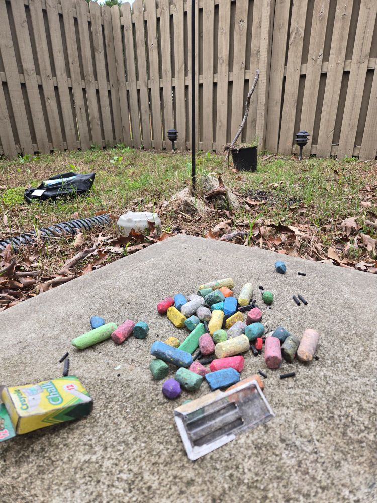 A pile of colored chalk sits on the corner of a concrete patio. Fallen leaves and grass stretch towards a wood fence in the background