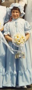 A rather dazed looking little girl in a pale blue bridesmaid’s dress with matching hat. She’s carrying a posey of daises