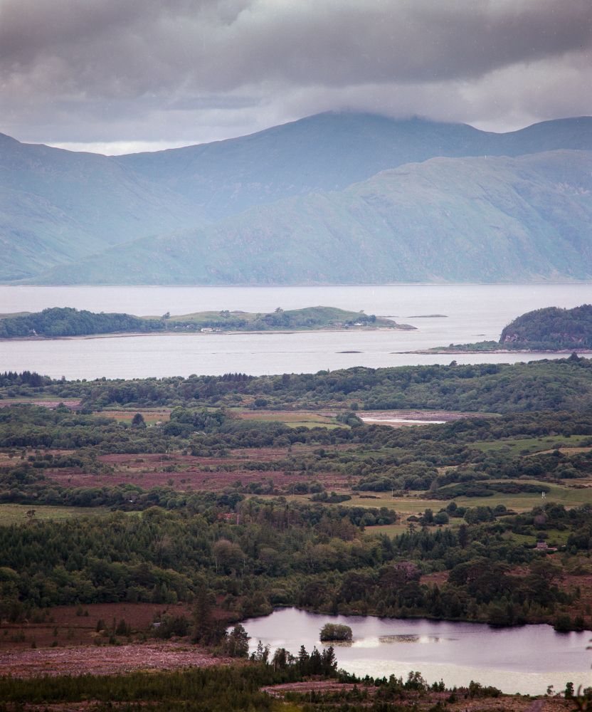 A layered landscape photograph taken on Kodak Ektar 100 film with a Mamiya RB67, capturing a moody, overcast scene in the Scottish Highlands. In the foreground, a small reflective loch is surrounded by dense woodland and moorland. The midground features lush green fields and rolling hills, leading to a wide estuary or sea loch. In the distance, towering mist-covered mountains rise under a dramatic sky heavy with dark clouds.