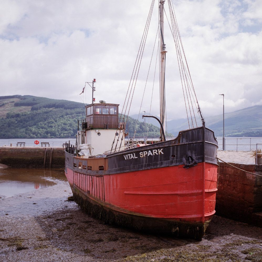 A vintage red and black puffer boat named "Vital Spark" sits on the muddy shore at low tide in Inveraray, Scotland. The boat is moored beside a stone quay, with green hills and calm waters of Loch Fyne in the background under a cloudy sky. The image was shot on Kodak Gold film using a Hasselblad camera, giving it a warm, nostalgic tone and fine detail.