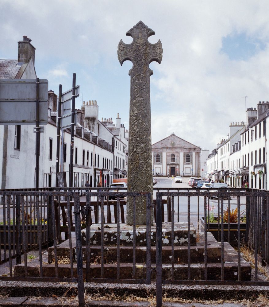 Photograph taken on Kodak Gold film with a Hasselblad camera, showing an intricately carved stone Celtic cross monument in the town centre of Inveraray, Scotland. The cross is framed by iron railings in the foreground, with white Georgian-style buildings lining both sides of the street and a neoclassical church at the end of the road under a partly cloudy sky.