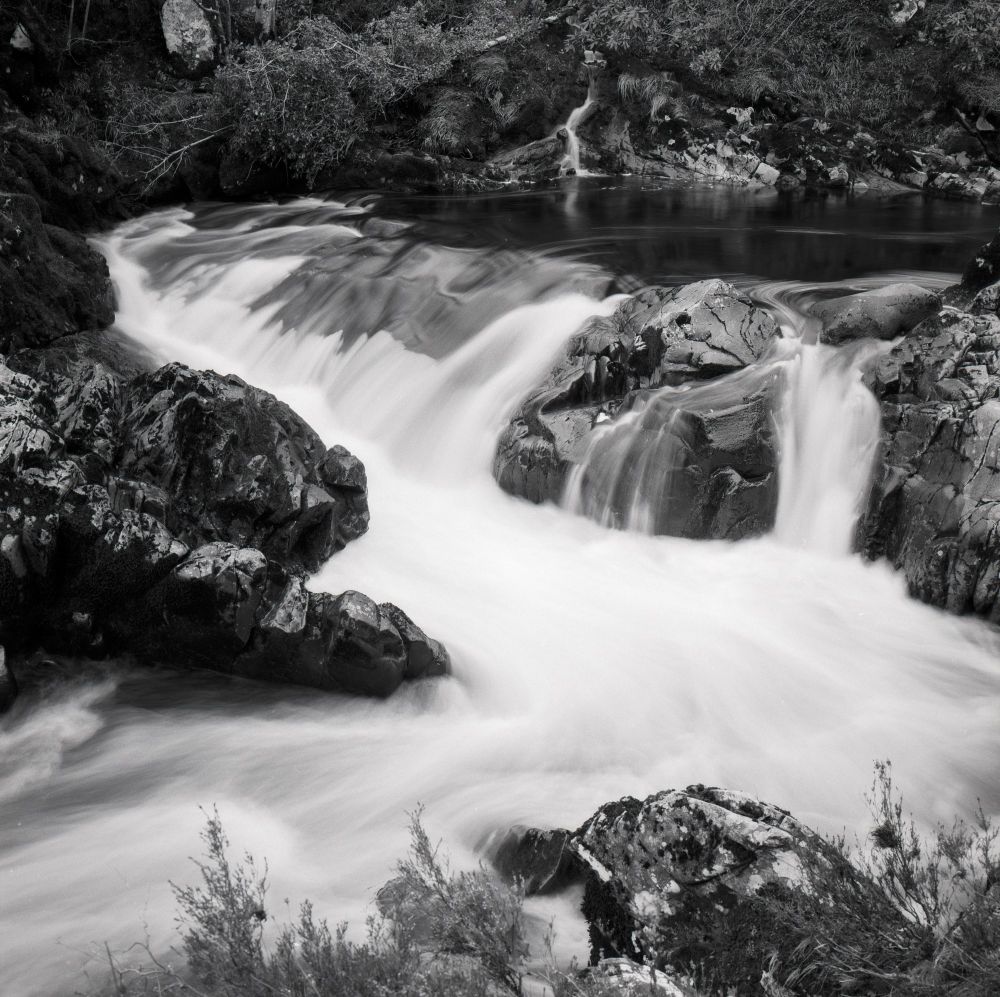 Black-and-white long exposure photograph of a small waterfall flowing over rugged rocks in Galloway Forest, Scotland, taken on Kodak TMax film with a Hasselblad camera. The smooth, silky water contrasts with the sharp textures of the rocks and surrounding vegetation.