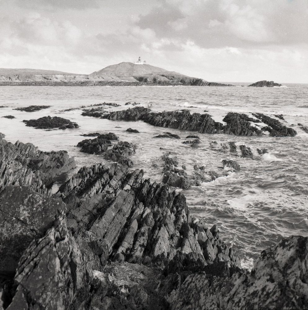 Black and white photograph of a rugged Irish coastline, with sharp, angular rock formations in the foreground and choppy waves crashing against them. In the distance, a small island with a lighthouse stands under a dramatic, cloud-filled sky. Shot on Kodak TMax film with a Hasselblad camera.