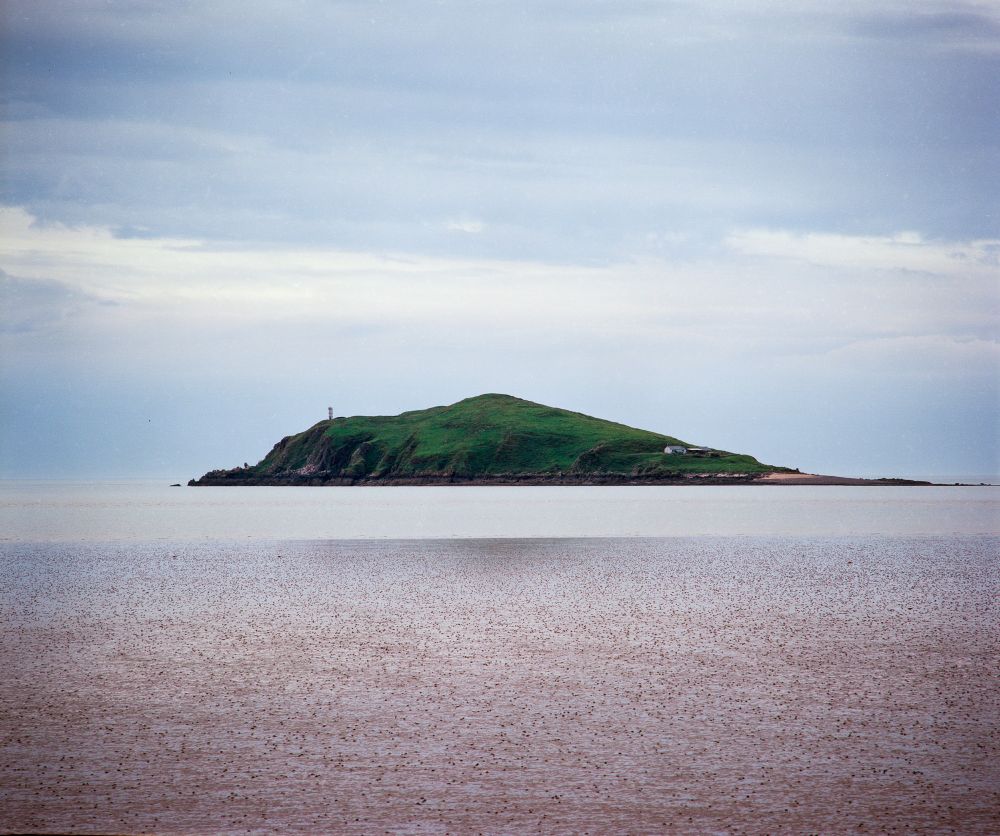 A distant view of a small, grassy island surrounded by calm tidal waters under a soft, overcast sky. Shot on Kodak Ektar 100 film using a Mamiya RB67 camera, somewhere along the Scottish coast.