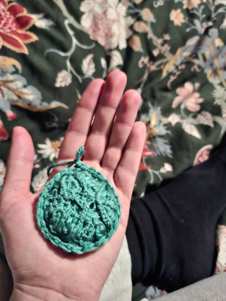 Round, crocheted shape. Lots of different braids and shapes of yarn, forming a turquoise amulet or medallion.  Placed in palm of hand. 