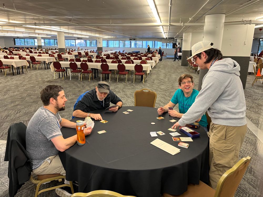 Three people seated at a round table playing card game, Alex standing and teaching, in a mostly-empty meeting hall with many tables and chairs in the background 