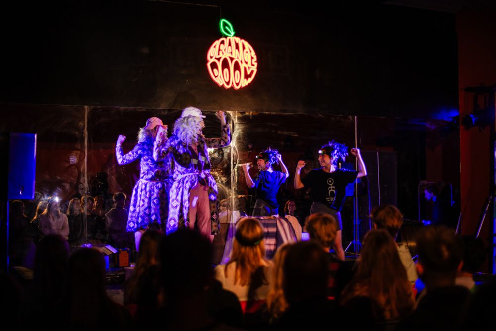 A drag queen in a house-coat flexes her muscles, encouraging a member of the audience to do the same.