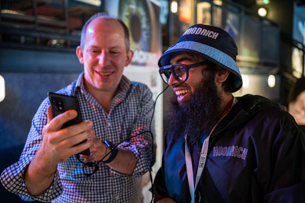 A man in a bucket hat wears a pair of eye-tracking glasses connected to a mobile phone being held by a second man.