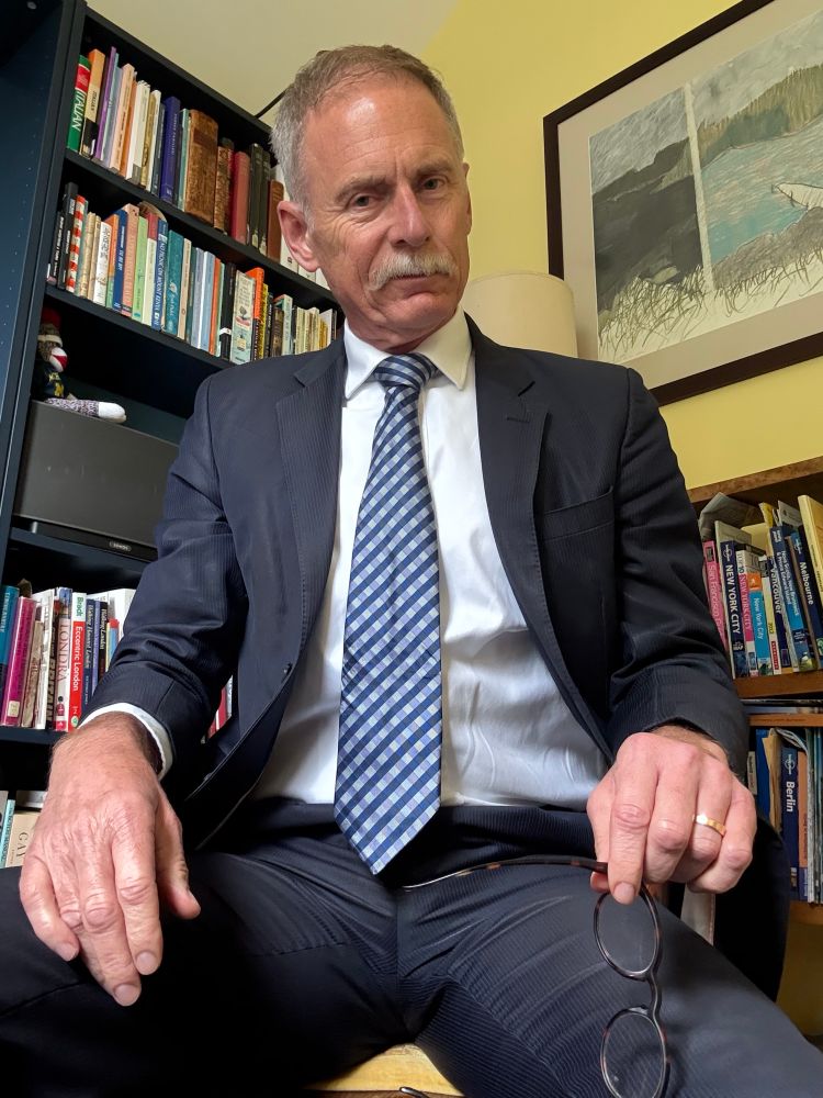 Andrew sitting suited with a blue patterned tie just off cam after a business meeting , glasses in hand