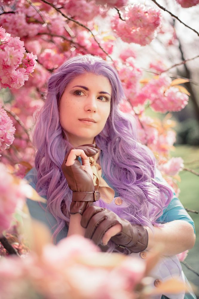 Purple haired woman surrounded by pink cherry blossom blooms. She is adjusting her glove and looking off to one side in thought. The character being portrayed is Imogen Temult from Critical Role.