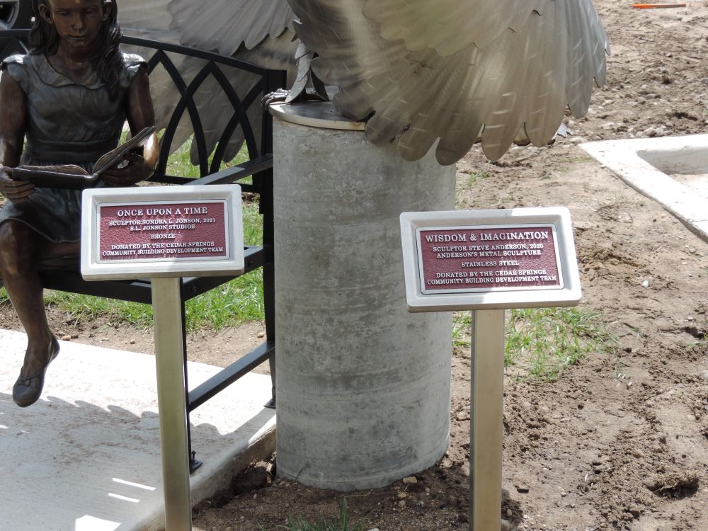 Two small plaques identifying the sculptures of "Once Upon a Time" and "Wisdom & Imagination" outside of the Cedar Springs library.