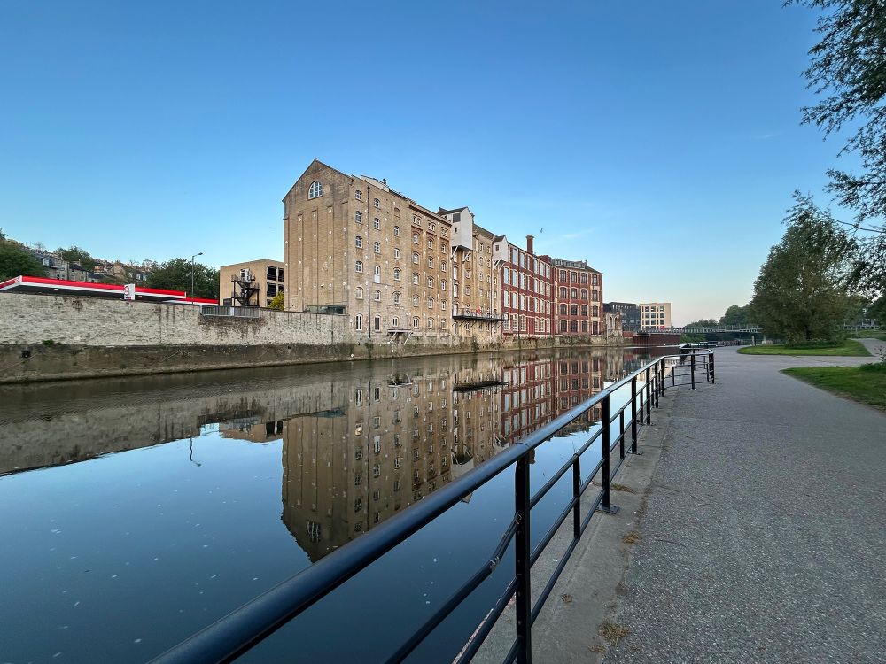 Early morning scene at the riverside. Former industrial buildings are reflected in the unusually calm and smooth River Avon. Camden Mill was a steam-powered flour mill and the red-brick building was a corset factory. Both are now office buildings. Bath, UK. Photo by Gearóid Burke. 