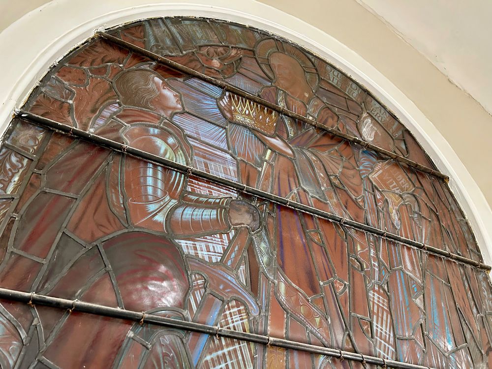 The top half of a stained glass window, as seen from below. The galleries of the main chapel now form part of the shop and the windows have been preserved (between the bookshelves). The glass is no longer backlit and limited light gives the artwork a brown cast, but some colour is still discernible. This scene depicts a figure in armour, presumably the archangel Michael, looking up to Christ, who is presenting them with a crown. At the top is the bible quote "Well done, thou good and faithful servant." 89 Broad Street, Reading, Berkshire, UK. Photo by Gearóid Burke.