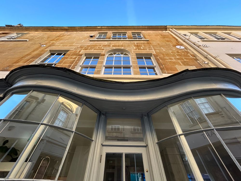 Outside a shop, looking up. The building has elegant undulating bow fronts at street level. Above, the more-typically Georgian windows do not align with the center of the shopfront. Which seems odd. Old Bond Street, Bath, UK. Photo by Gearóid Burke.